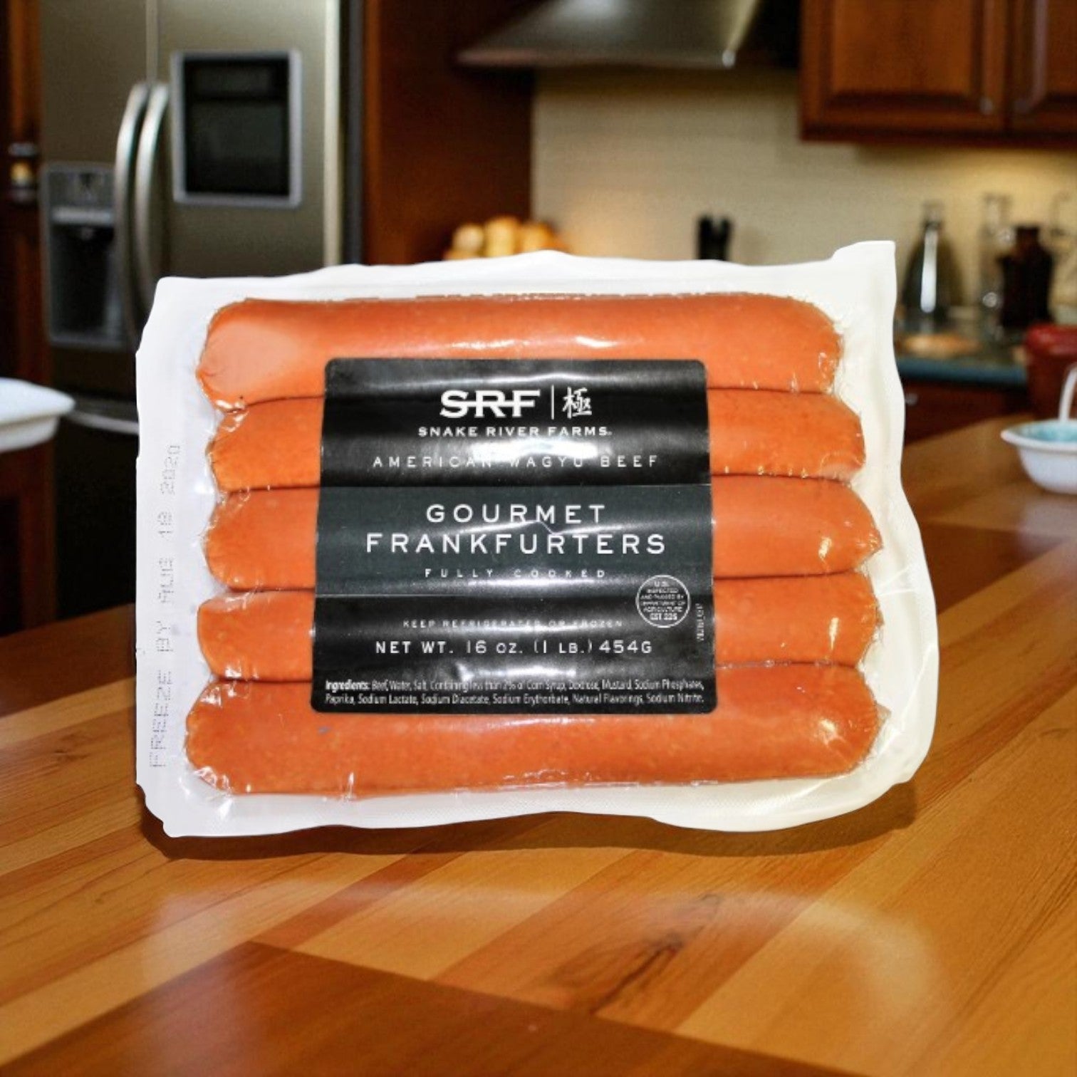 A vacuum-sealed package of Snake River Farms Wagyu Hot Dogs - 1 lb rests on a wooden counter, with a stainless steel fridge and brown cabinets behind—ideal for creating a gourmet hot dog experience.