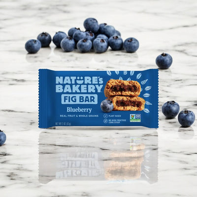 A package of Nature's Bakery Blueberry Fig Bar - 2 oz sits on a marble surface with scattered blueberries around it. The packaging highlights that these plant-based bars from Nature's Bakery are non-GMO, gluten-free, and contain real fruit and whole grains.