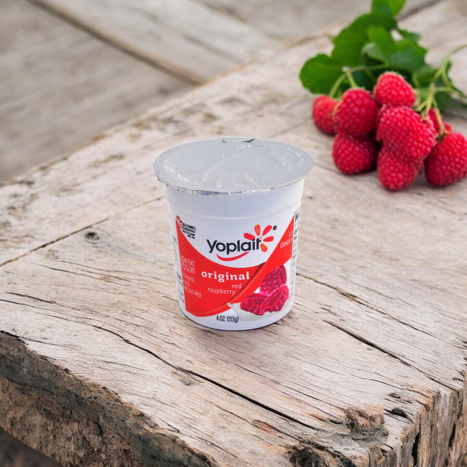 A Yoplait Original Raspberry and Peach Yogurt cup, celebrated for its creamy texture and lack of artificial flavors, rests on a rustic wooden surface with fresh raspberries and green leaves in the background.