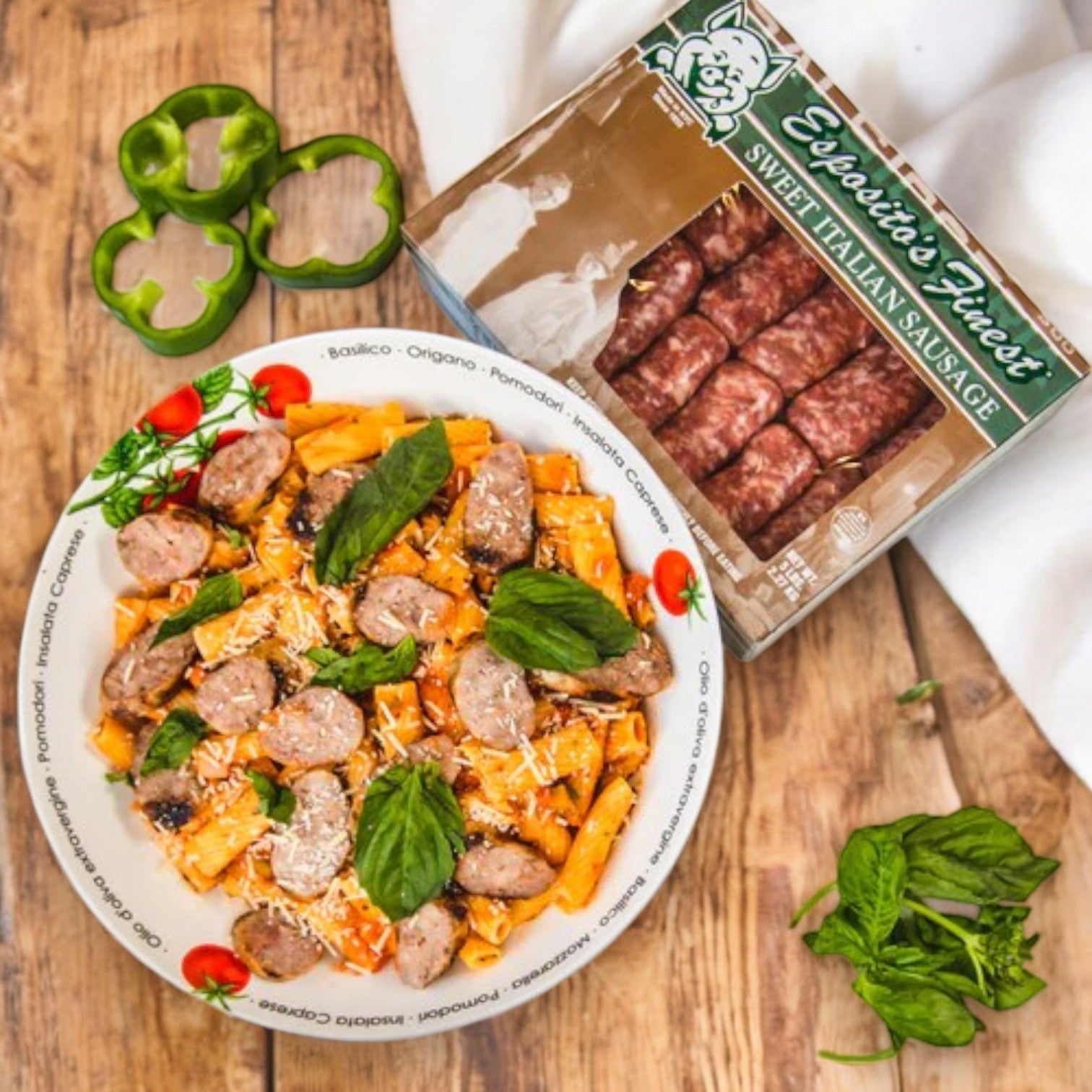 A bowl of pasta with sausage slices and basil sits on a wooden table, exuding authentic Italian flavor. Next to it is a package of Esposito's Finest Quality Sausage – Sweet Italian Sausage, alongside sliced green bell peppers, showcasing the premium ingredients in this delightful dish.