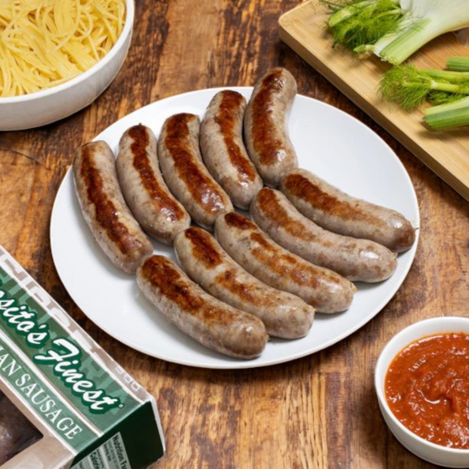 A plate of Esposito's Finest Quality Sweet Italian Sausage on a wooden table is surrounded by uncooked pasta, tomato sauce, and fresh fennel. These premium sausages ensure an authentic Italian flavor in every bite.